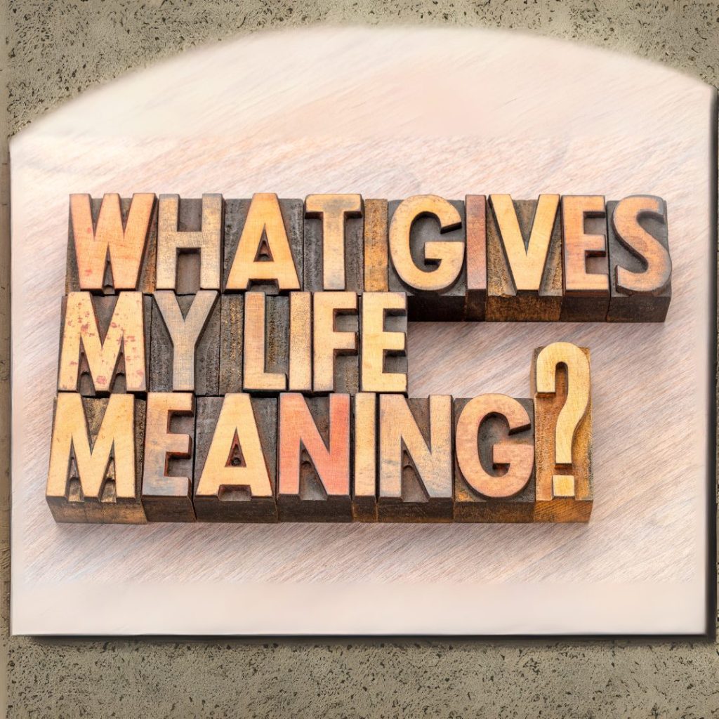 Wooden letter blocks spelling out "WHAT GIVES MY LIFE MEANING?" arranged on a light-colored wooden surface. The letters are in different shades of brown, orange, and cream, with some showing signs of wear and age.