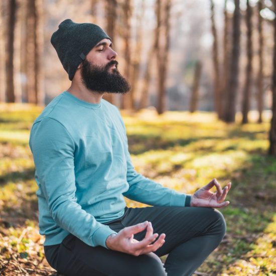Man wearing a beanie and a long shirt is sitting down outside meditating.