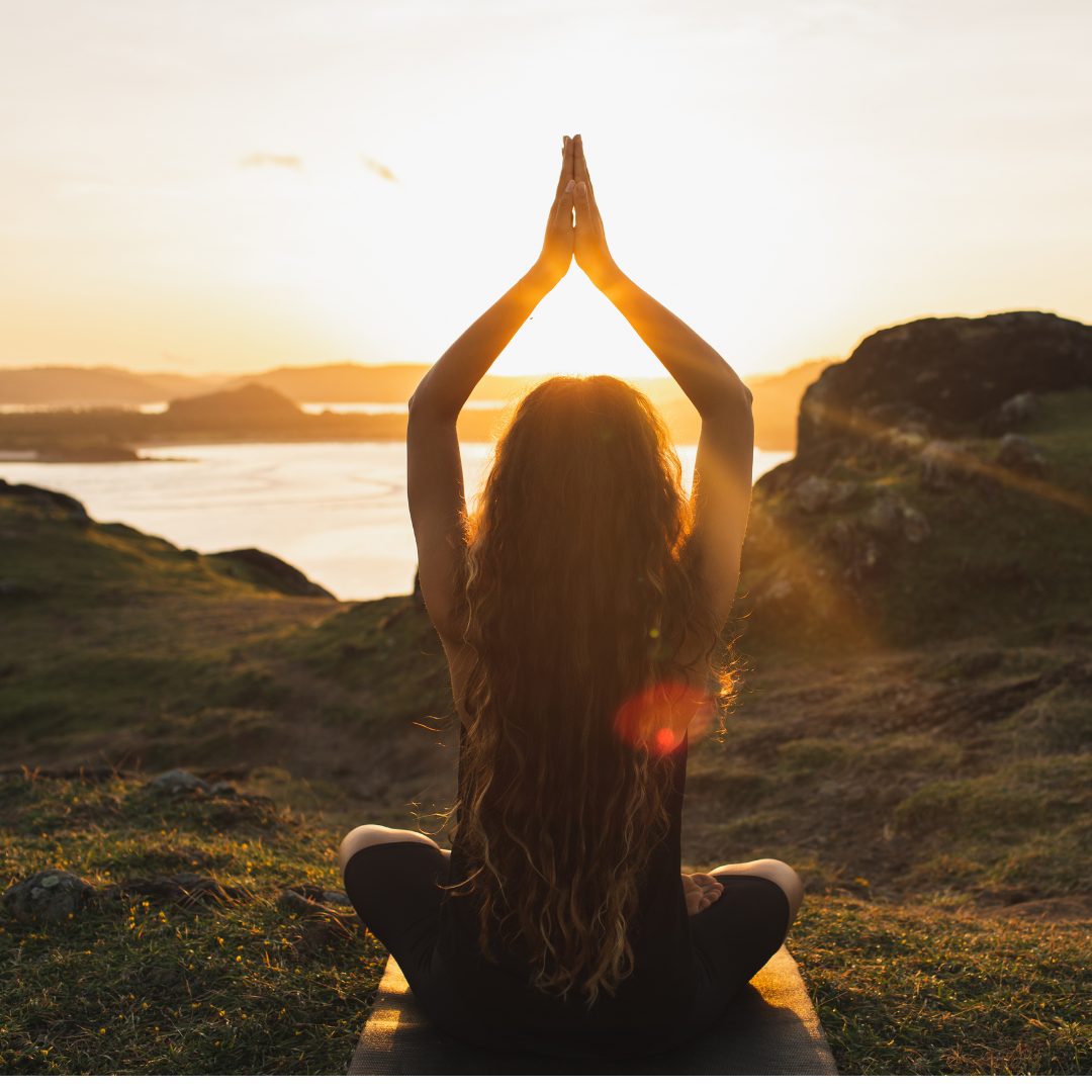 A woman with long, wavy hair is sitting cross-legged on a yoga mat outdoors during sunset. She is facing away from the camera with her hands pressed together overhead in a prayer position. The scene features a scenic landscape with rolling hills, a body of water, and the sun setting in the background, casting a warm golden glow.