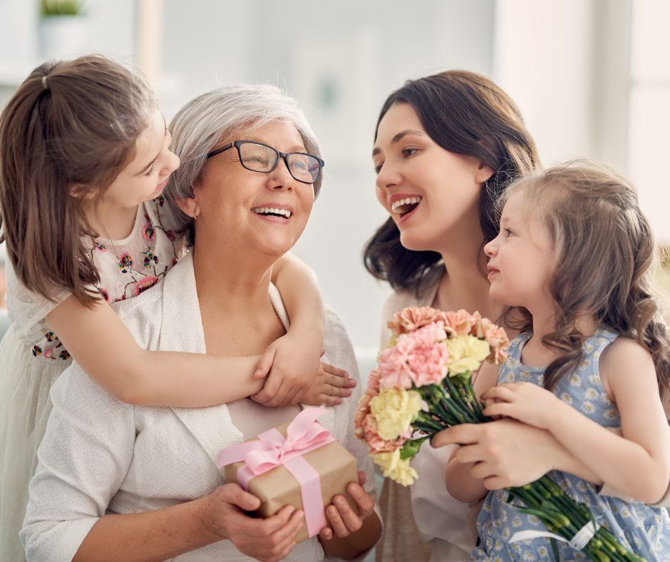 A joyful scene of three generations of women celebrating together. An elderly woman with gray hair and glasses is smiling happily, holding a gift wrapped with a pink ribbon. A young woman with dark hair is leaning in close, smiling warmly at the elderly woman. Two young girls, one holding a bouquet of pink and yellow flowers and the other hugging the elderly woman, are also smiling and sharing the moment. The background is bright and softly lit.