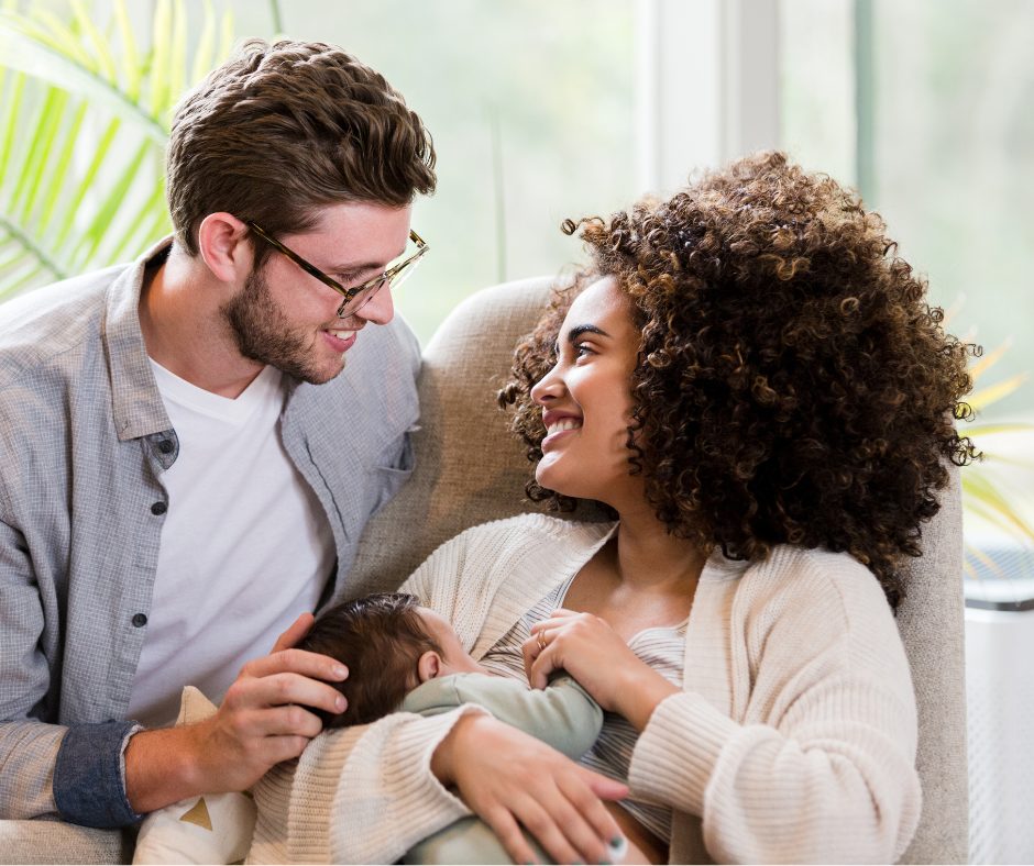 A couple sitting comfortably on a sofa, sharing a tender moment as they look at each other. The woman is breastfeeding a baby, and both are smiling warmly. The background shows a bright room with large windows and green plants.