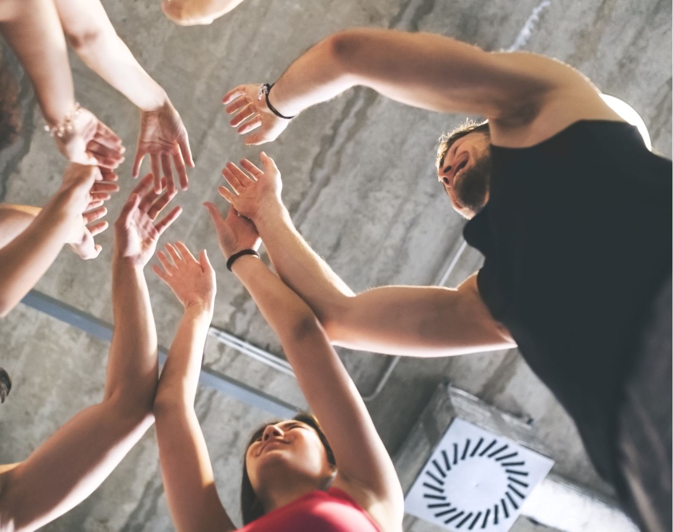 A group of people reaching their hands upward toward a man with a beard and muscular arms, who is leaning over and smiling. The photo is taken from below, showing the participants' hands and faces against a concrete ceiling with a ventilation grate visible in the background.