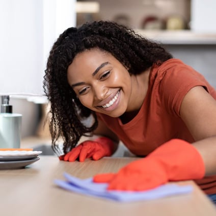 A woman with curly hair smiling happily as she cleans a wooden table. She is wearing an orange t-shirt and orange cleaning gloves, and appears to be enjoying her task in a bright, modern kitchen.