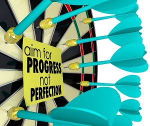 Dartboard filled with missed darts surrounding a sticky note that reads “aim for PROGRESS not PERFECTION,” emphasizing growth over flawlessness.