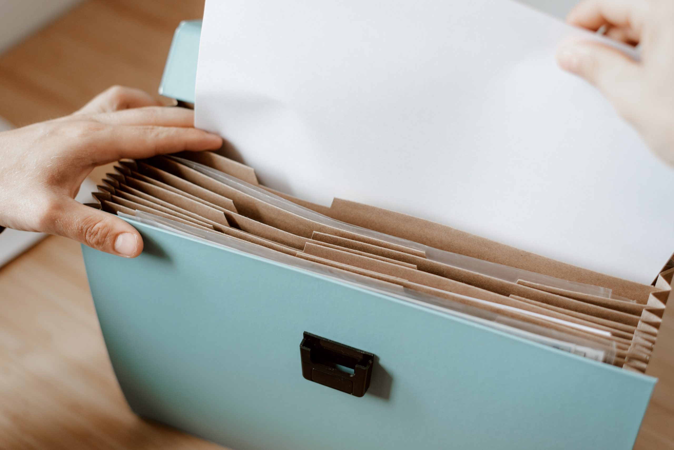 The image shows a person placing a white sheet of paper into a light blue filing box that contains multiple hanging file folders. The box is on a wooden surface, and the person's hands are visible, one holding the paper and the other opening the file.