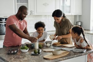 A family is gathered in a bright, modern kitchen, preparing food together. An adult man in a pink shirt is spreading something onto bread, while a woman in a beige blouse is holding a slice of bread and smiling. Two children, a boy with curly hair and a girl with braided hair, are watching and participating; the boy is eating, and the girl is leaning on the counter. The kitchen island has various ingredients, bowls, and cutting boards, creating a warm, collaborative atmosphere.