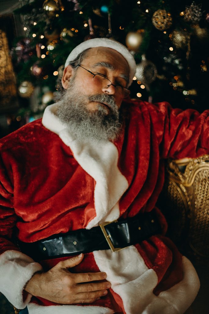 The image shows a man dressed as Santa Claus resting with his eyes closed, sitting in front of a decorated Christmas tree with glowing lights and ornaments. He is wearing a traditional red velvet Santa suit with white fur trim, a black belt, and a Santa hat. His hand is resting on his stomach, and he has a long gray beard, glasses, and a peaceful expression. The background features warm, festive holiday decor.