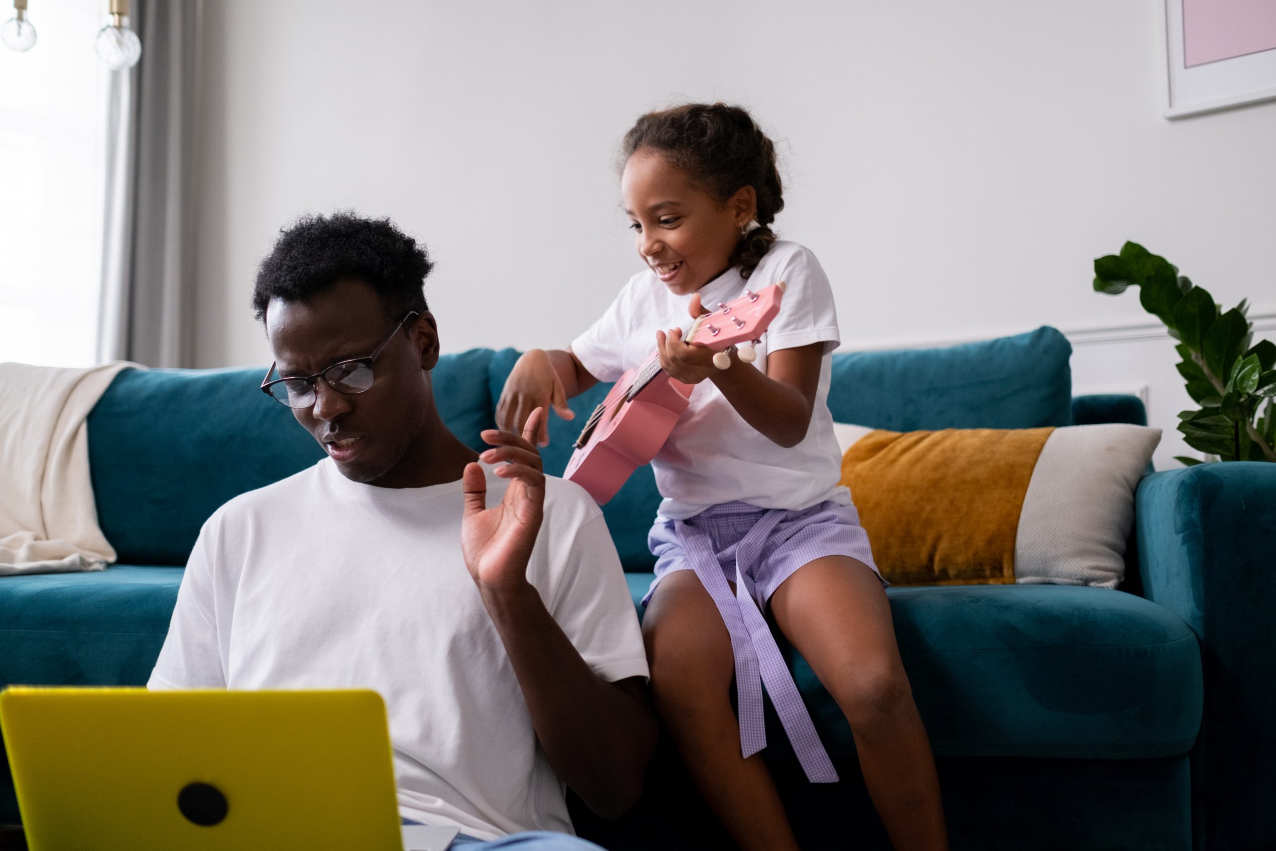 The image shows a young girl sitting on a teal sofa, playing a pink ukulele and smiling joyfully. In front of her, a man wearing glasses and a white t-shirt is sitting cross-legged on the floor, looking at a yellow laptop with a focused expression. The background features a modern living room with a beige blanket draped over the sofa and decorative pillows, with natural light coming in from a window on the left.