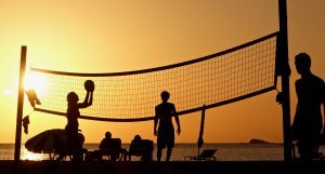 Silhouettes of people playing volleyball on the beach during sunset, with the ocean and a small island in the background. The sky is a warm orange hue, and some beach chairs and an umbrella are visible near the shoreline.