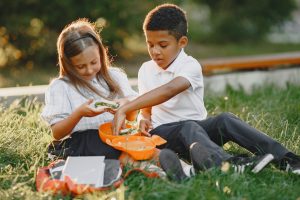 A young girl and a boy are sitting on the grass outdoors, playing with a toy fishing set. The girl is holding a toy fish, and the boy is reaching into an orange toy container. They are surrounded by a few toys and a small chalkboard, with trees and greenery in the background, enjoying a sunny day outside.
