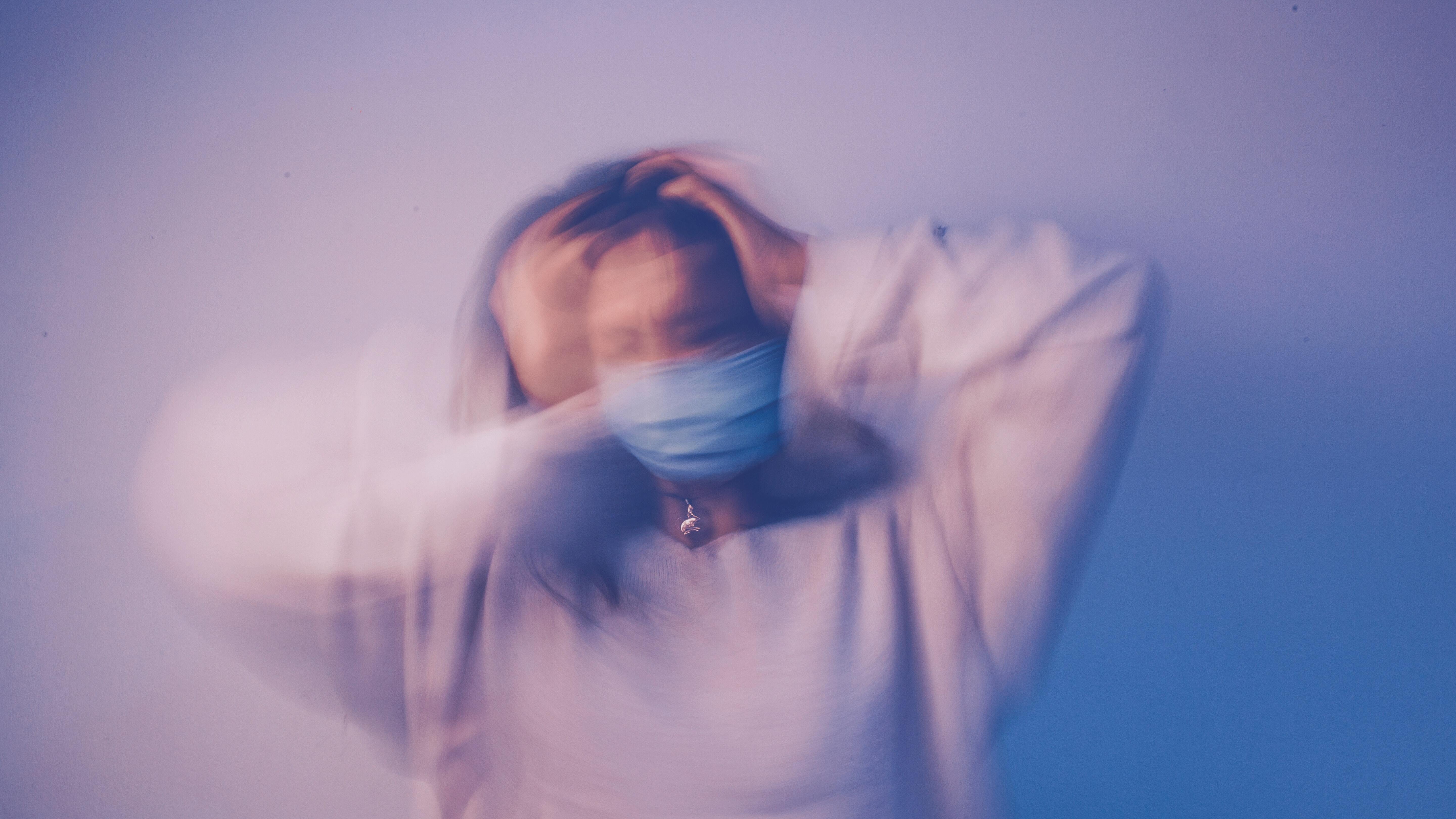 Blurred image of a woman wearing a face mask, holding her head with both hands, symbolizing anxiety, stress, or emotional overwhelm.