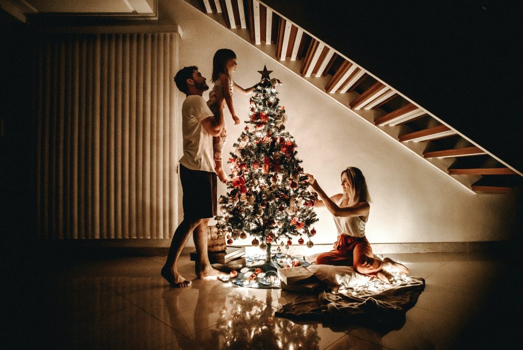 The image shows a family decorating a Christmas tree indoors at night. A man is standing and holding a young girl on his shoulders, who is reaching out to place an ornament on the tree. A woman is kneeling on a blanket on the floor, smiling and helping to decorate the tree. The scene is warmly lit by the lights on the Christmas tree, creating a cozy and joyful holiday atmosphere underneath a staircase.