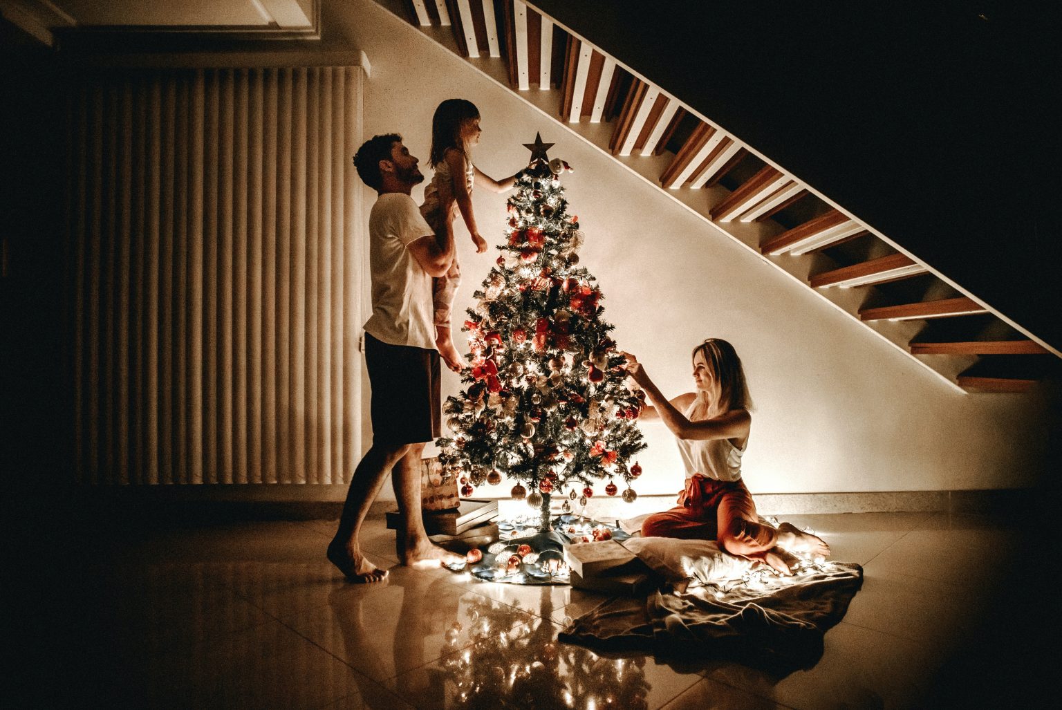 The image shows a family decorating a Christmas tree indoors at night. A man is standing and holding a young girl on his shoulders, who is reaching out to place an ornament on the tree. A woman is kneeling on a blanket on the floor, smiling and helping to decorate the tree. The scene is warmly lit by the lights on the Christmas tree, creating a cozy and joyful holiday atmosphere underneath a staircase.