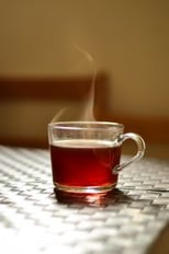 A clear glass cup filled with hot coffee, with steam rising from the surface. The cup is placed on a white, textured surface, and the background is softly blurred with warm, neutral tones.