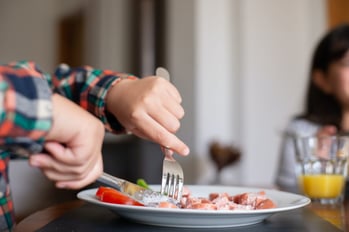 A person wearing a colorful plaid shirt is cutting raw meat on a white plate with a fork and knife. In the background, there is a glass of orange juice and a blurred woman with dark hair. The scene appears to be a meal preparation in a home setting.