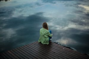 Girl sitting alone at the edge of a wooden dock, looking out over calm, reflective water, conveying solitude and deep contemplation.