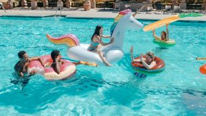 A group of five people enjoying a sunny day in a swimming pool with inflatable floats. One woman is sitting on a large, white unicorn-shaped float, while others are on various smaller floats, including a pink donut-shaped float, an orange donut-shaped float, and a yellow float with a woman holding a large inflatable ring above her head. The scene appears fun and lively, with everyone smiling and engaging with each other.