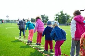 A group of young children, dressed in colorful jackets and pants, are participating in an outdoor activity on a grassy sports field. They are stepping through a series of small ladders laid out on the ground, supervised by an adult. The background features trees, a water tower, and a clear sky, indicating a bright, sunny day.