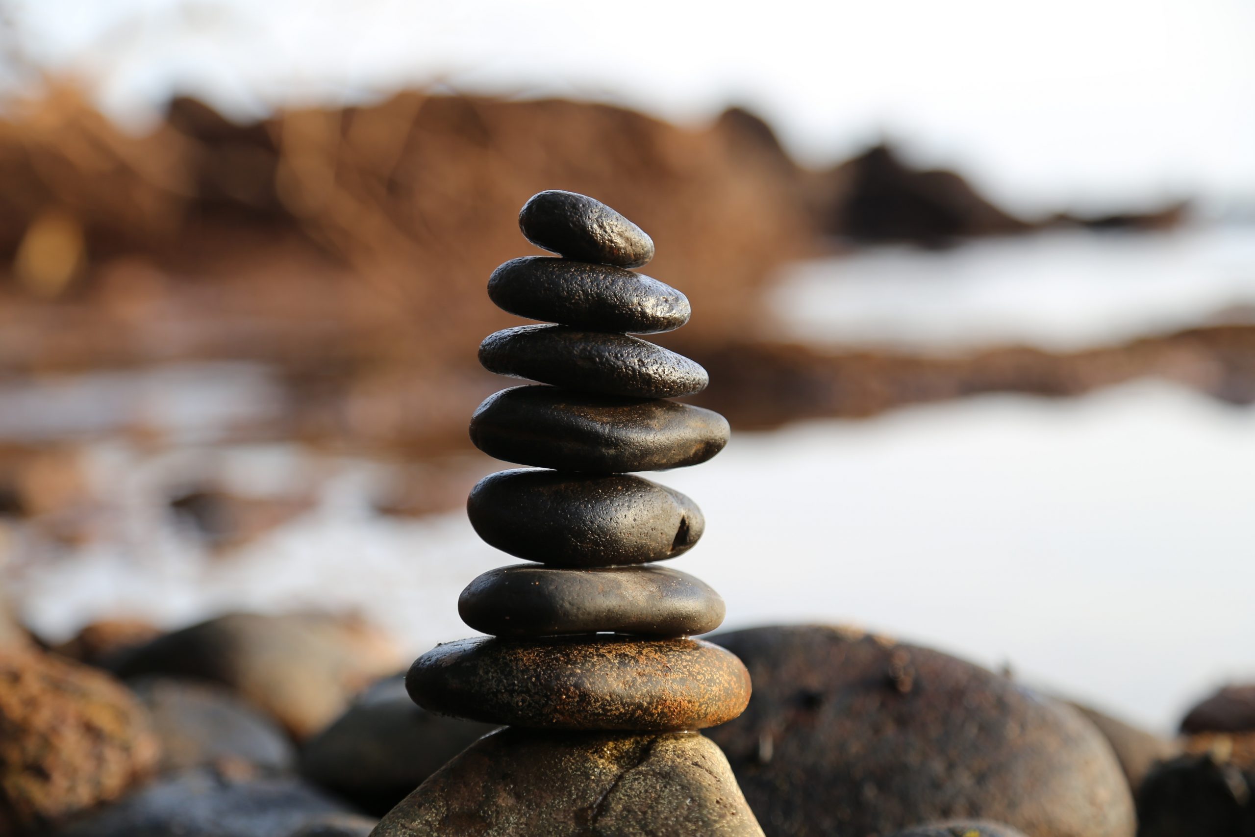 The image shows a vertical stack of smooth, dark stones balanced on top of each other on a rocky surface, with a blurred background of more rocks and water.