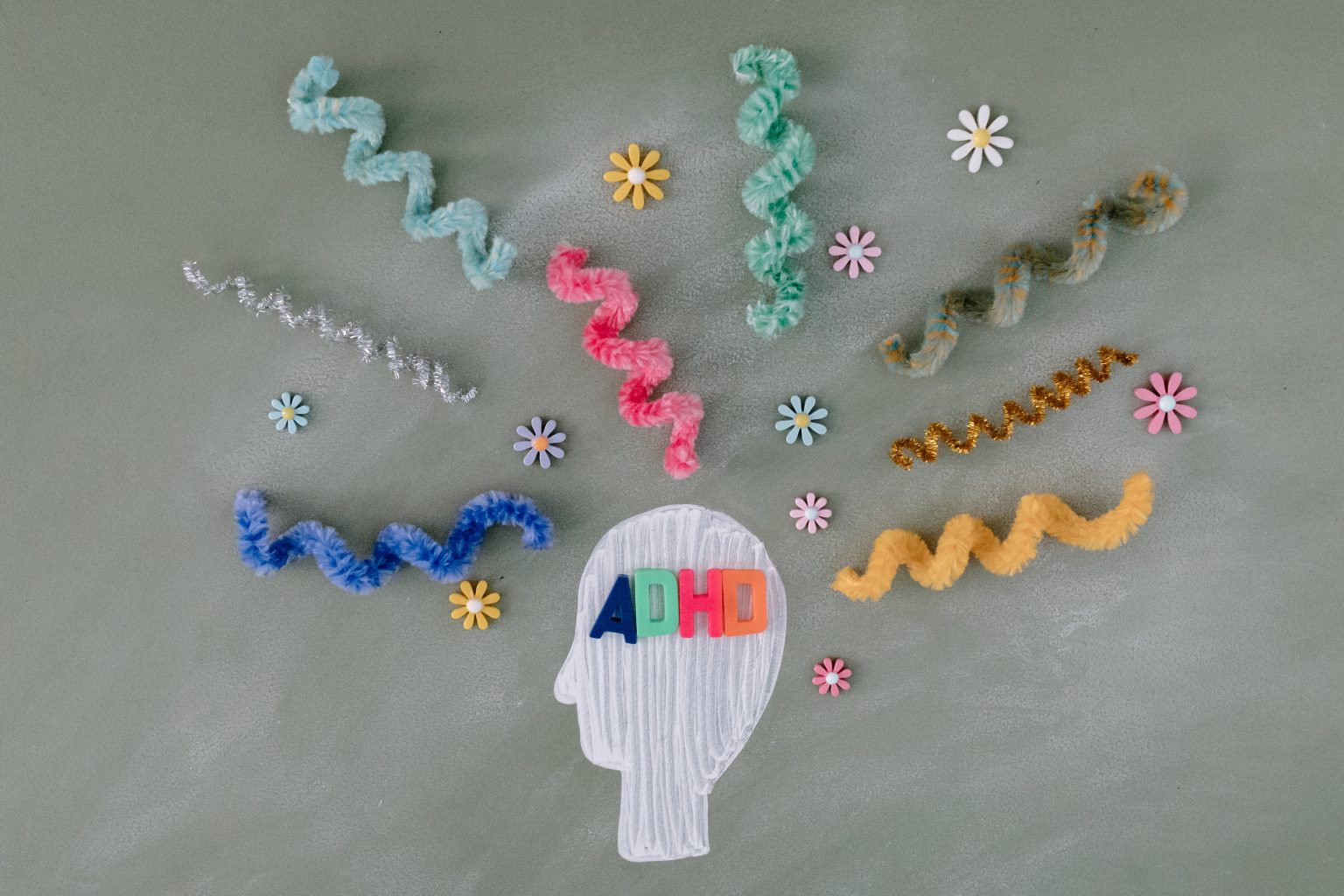 A paper cutout of a human head in profile view, with colorful foam letters spelling "ADHD" inside the head, surrounded by various decorative pipe cleaners and small flower-shaped embellishments on a neutral background.
