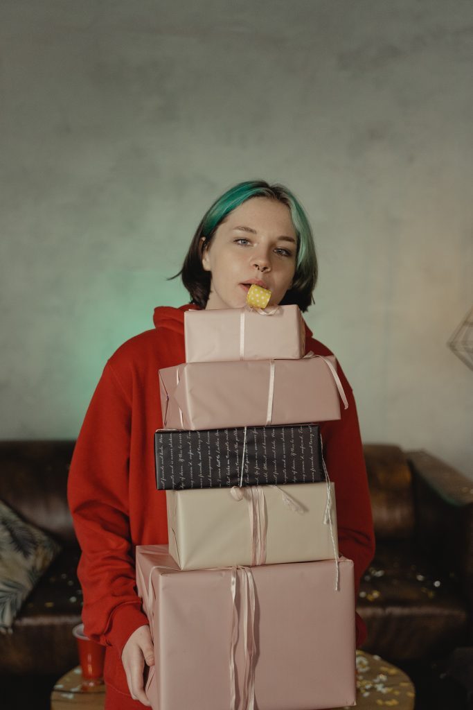 The image shows a young woman with short, teal-colored hair standing indoors, holding a tall stack of wrapped presents. She is wearing a red hoodie and has a playful expression, with a yellow gift tag in her mouth. The background features a plain, light-colored wall and a brown leather sofa. There are some scattered confetti or small decorative items on the table in front of her.