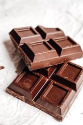 A close-up of a broken milk chocolate bar with square segments, resting on crumpled white paper. The chocolate has a smooth surface with a shiny finish, and some pieces are slightly chipped or broken.