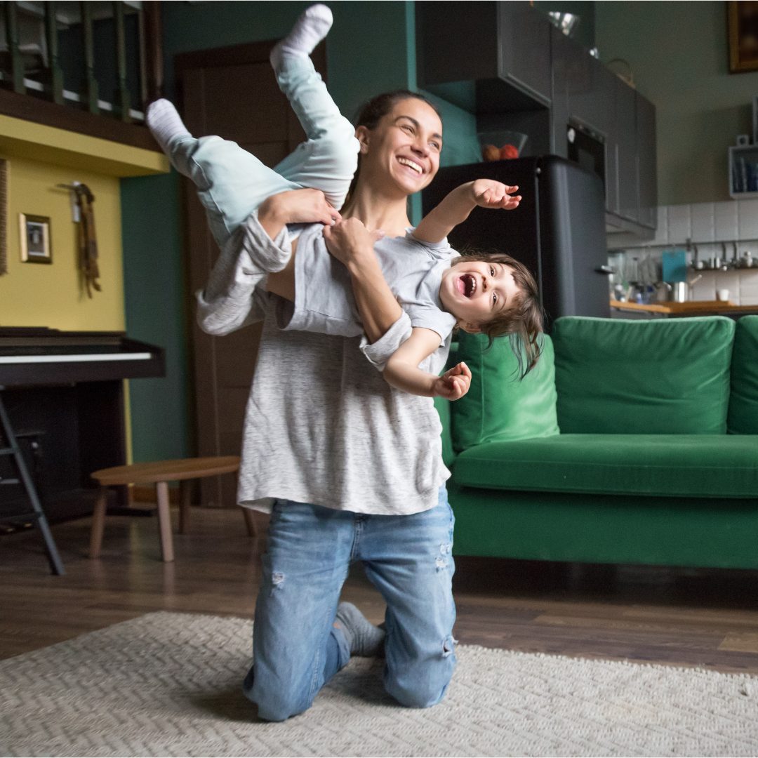 A woman is kneeling on the floor, smiling happily while holding a young girl upside down in her arms. The girl has a big, joyful expression on her face, with her mouth open as if laughing, and her arms extended outward. They are in a cozy living room with a green couch, a piano, and kitchen appliances visible in the background.