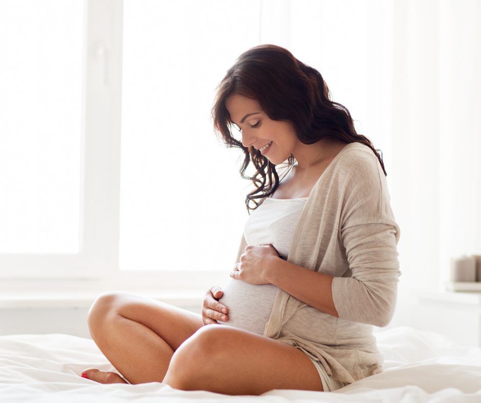 A pregnant woman sitting on a bed, smiling happily and gently holding her baby bump with both hands. She has dark, wavy hair and is wearing a light-colored top and cardigan, with a bright, softly lit background.
