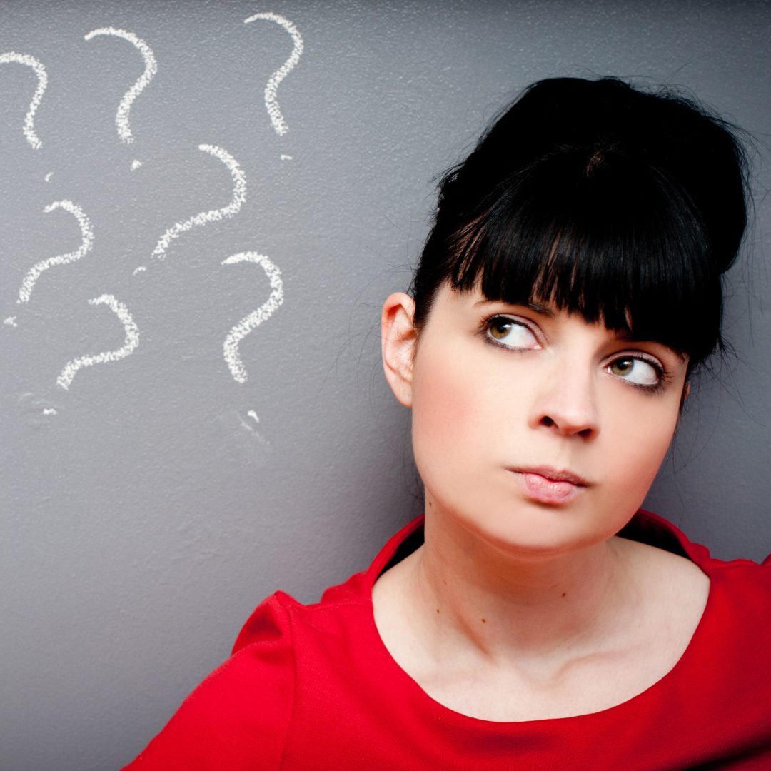 A woman with dark hair styled in an updo and bangs is looking slightly upward and to the side with a thoughtful expression. She is wearing a red top and is standing against a gray wall. Several white question marks are drawn on the wall to her left, indicating confusion or pondering.