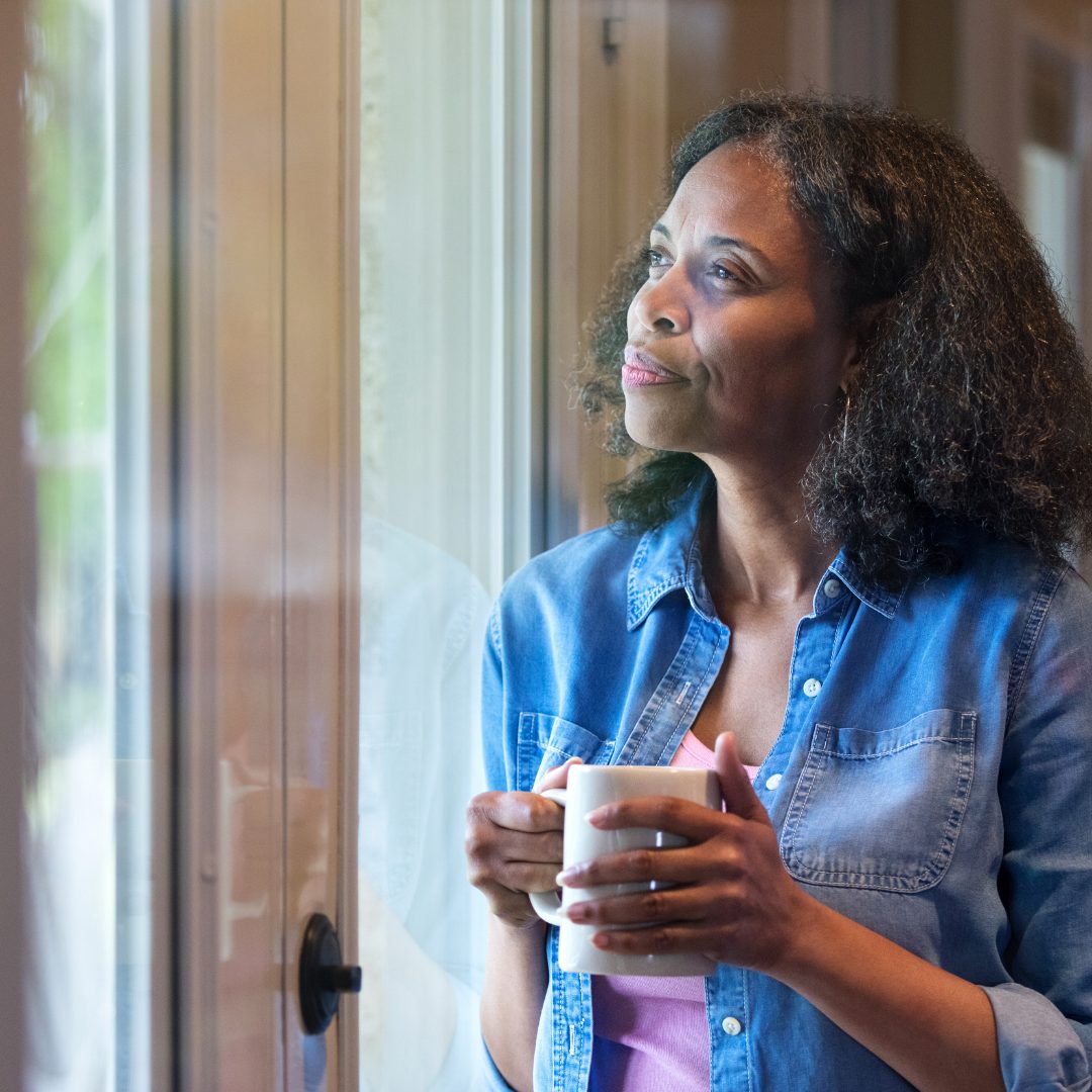 A woman with curly dark hair and medium skin tone is standing by a window, holding a white mug in both hands. She is wearing a denim shirt over a pink top and gazes thoughtfully outside with a serene expression, enjoying a quiet moment. The background shows part of a cozy indoor setting with warm lighting.