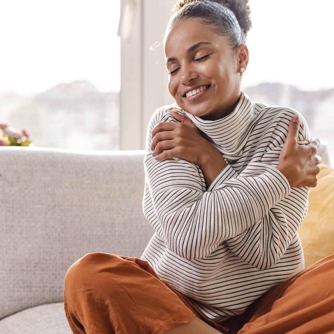 A young woman with curly hair, smiling softly with her eyes closed, hugging herself with both arms crossed over her chest. She is sitting on a light-colored sofa in a bright room, with a large window behind her letting in natural light. She appears to be feeling content and peaceful.
