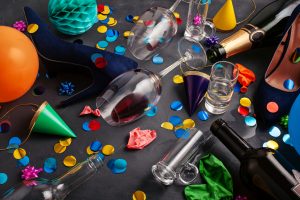 A cluttered celebration scene featuring empty and half-empty glasses, colorful confetti, party hats, balloons, crumpled paper, and a bottle of champagne. The table is covered with scattered multicolored paper circles and various party decorations, creating a lively and festive atmosphere.