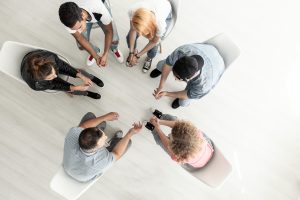 Six people sitting in a circle, holding hands and engaging in a group activity or prayer, viewed from above in a bright, minimalistic room with light-colored flooring and white chairs.