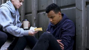 A young man in a gray hoodie sitting outside against a wooden fence, holding a coffee cup, with a sandwich on his lap and a serious expression.