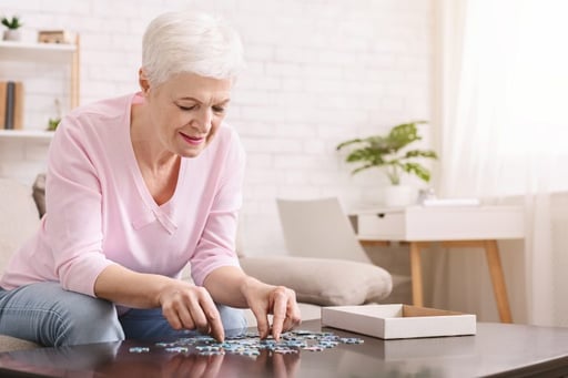 An elderly woman with short white hair is sitting at a table, working on assembling a jigsaw puzzle. She is wearing a light pink top and appears to be focused on fitting the pieces together. The room has a bright, cozy atmosphere with a white brick wall, a bookshelf, a potted plant, and a window with curtains in the background.