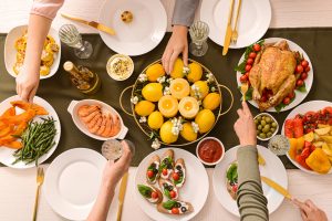 A top-down view of a table set for a celebration with various dishes and drinks. The table includes a roasted chicken with vegetables, a plate of smoked salmon, a bowl of olives, a platter of colorful vegetables, and a variety of appetizers. There are also glasses of wine, bottles of beer, and a bowl of lemon-colored desserts. Several hands are reaching for food, and the table is decorated with cutlery, napkins, and small bowls of sauces, creating a festive and communal atmosphere.