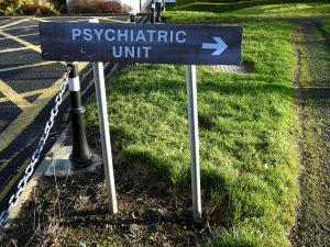 Outdoor sign reading “Psychiatric Unit” with an arrow pointing right, positioned along a grassy pathway.
