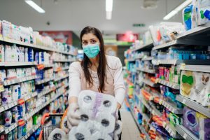 A woman wearing a blue face mask is shopping in a grocery store aisle, pushing a shopping cart and holding a pack of toilet paper.