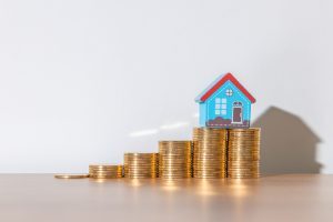 Small toy house sitting on top of ascending stacks of gold coins, symbolizing increasing home value or real estate investment growth.