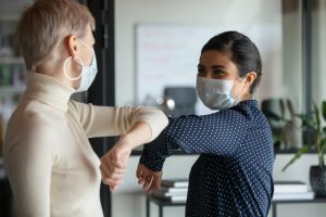 Two women wearing face masks are greeting each other with an elbow bump in an indoor setting, smiling and maintaining social distance.