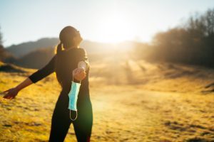 Woman standing in a sunlit field with arms outstretched, holding a face mask in one hand, expressing freedom and relief.