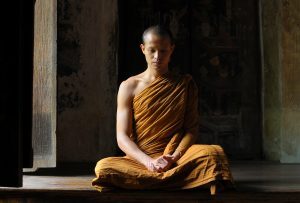 A young Buddhist monk sitting cross-legged on the floor, dressed in traditional orange robes, meditating in a dimly lit room with wooden walls.