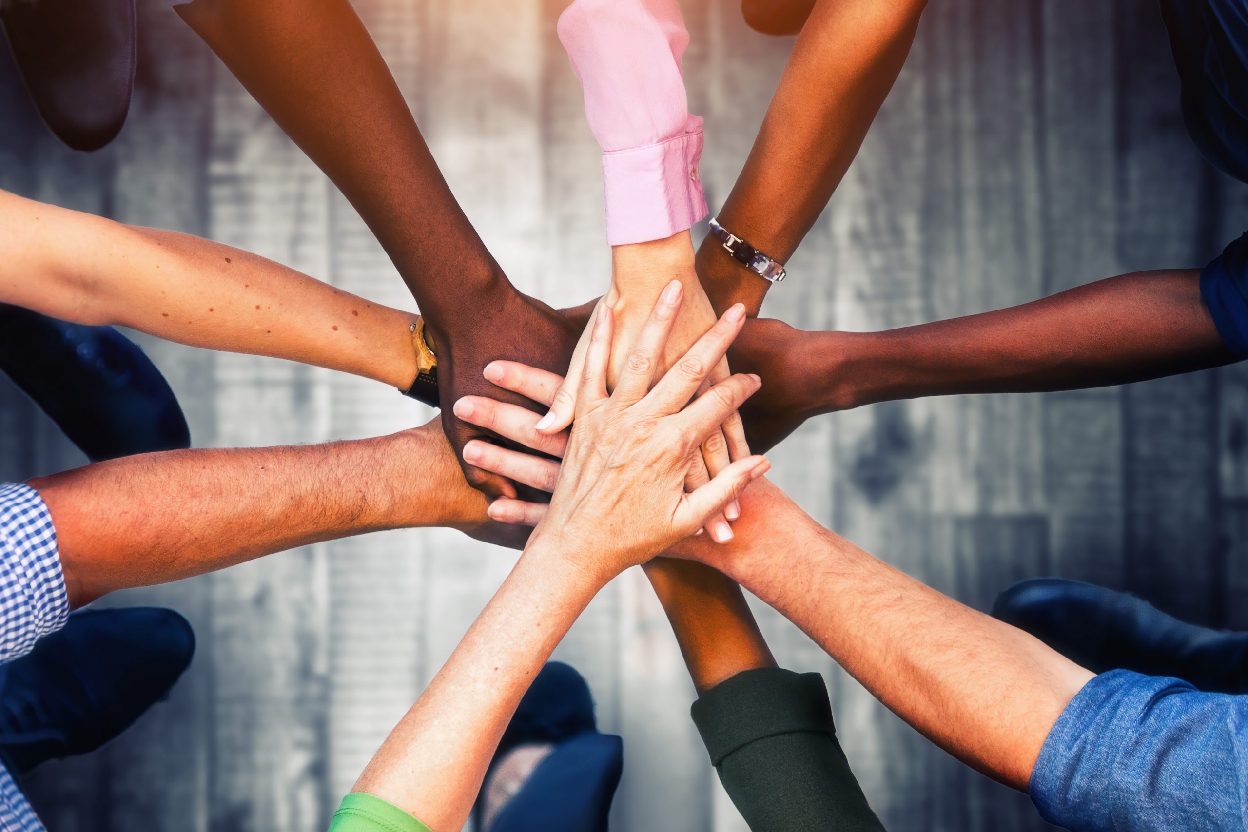 A group of diverse hands stacked together in the center, symbolizing unity, teamwork, and collaboration. The hands vary in skin tones and are reaching in from different directions, all meeting in a show of support and solidarity.