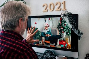 An elderly man with gray hair wearing a red plaid shirt waves at a computer screen displaying a woman in a Santa hat sitting in a decorated room with a Christmas tree, gifts, and holiday ornaments.