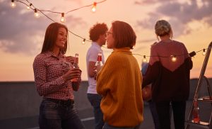 Group of friends socializing outdoors at sunset with string lights overhead, two women smiling and chatting while holding drinks.