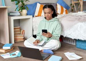 A young woman sitting on the floor of her bedroom, using a smartphone while a laptop and open books are nearby, surrounded by scattered papers and notebooks.
