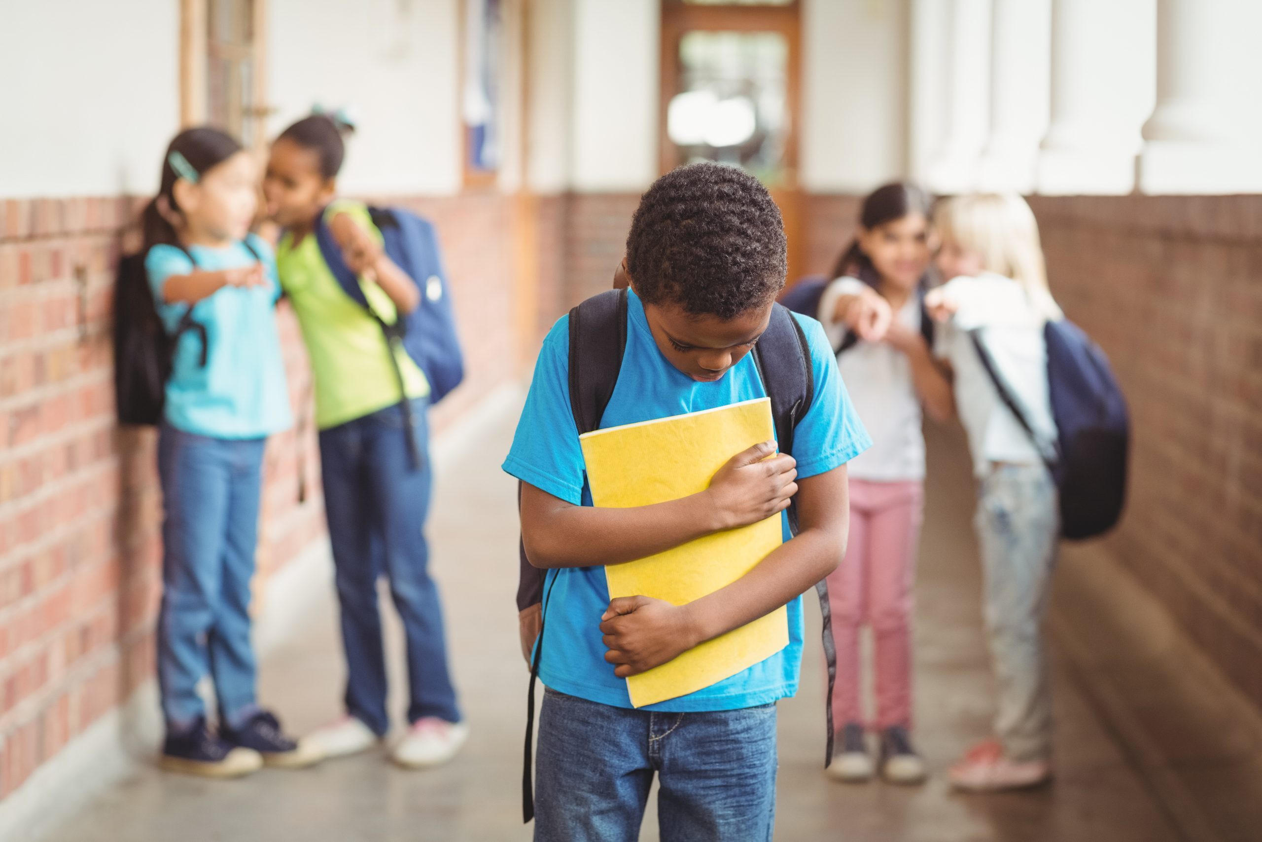 Several children standing and chatting in a school hallway, with one boy in the foreground holding a yellow folder and looking down, while others are in the background talking and smiling.