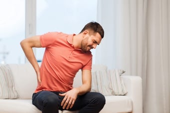 A man sitting on a white sofa, grimacing in pain and holding his lower back with one hand.