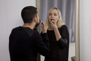 A woman with blonde hair is standing in front of a mirror, applying red lipstick. She is wearing a black top with leather sleeves. A man with dark hair and a beard, dressed in black, is standing close to her, holding a lipstick tube near her lips as he assists her. The background includes a gray curtain and a white wall.