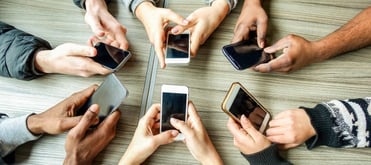 A group of six people sitting around a table, each holding a smartphone, with their hands and devices visible from above.
